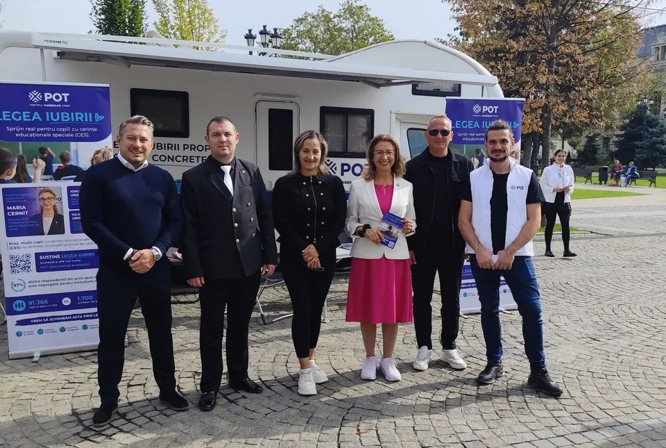 Șapte persoane stau în rând în fața unui vehicul alb pe care sunt afișate panouri informative colorate.