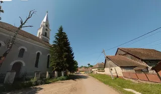 O clădire albă cu un turn înalt și un conac din lemn stau de o parte a unui drum de țară, sub un cer senin.