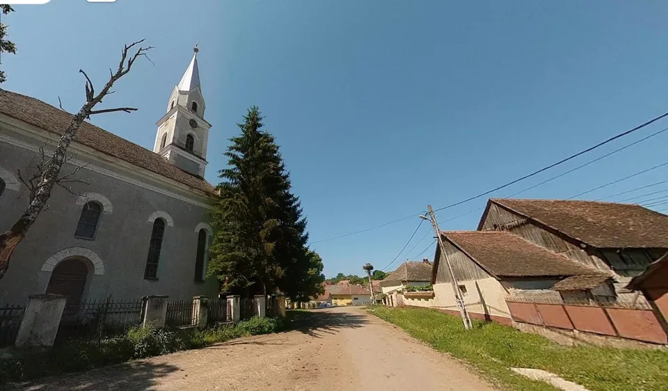 O clădire albă cu un turn înalt și un conac din lemn stau de o parte a unui drum de țară, sub un cer senin.