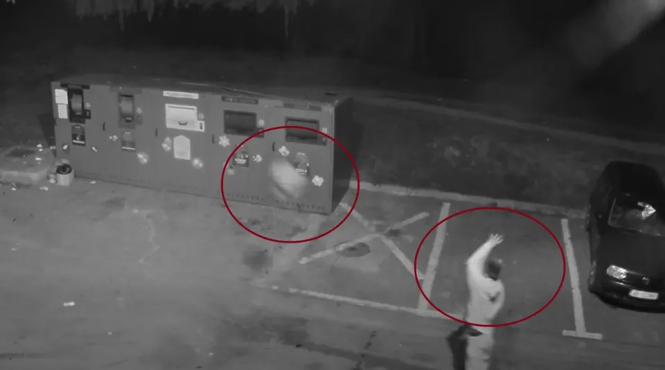 O imagine alb-negru de supraveghere surprinde două zone cerculate în roșu pe un teren asfaltat lângă un container mare și o mașină în parcarea de lângă acesta.