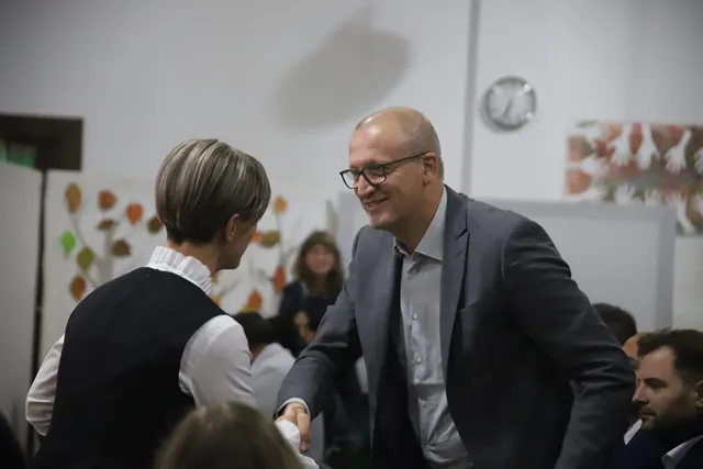 Un bărbat cu ochelari și sacou gri întinde mâna spre o femeie cu vestă neagră și cămașă albă într-o încăpere interioară.