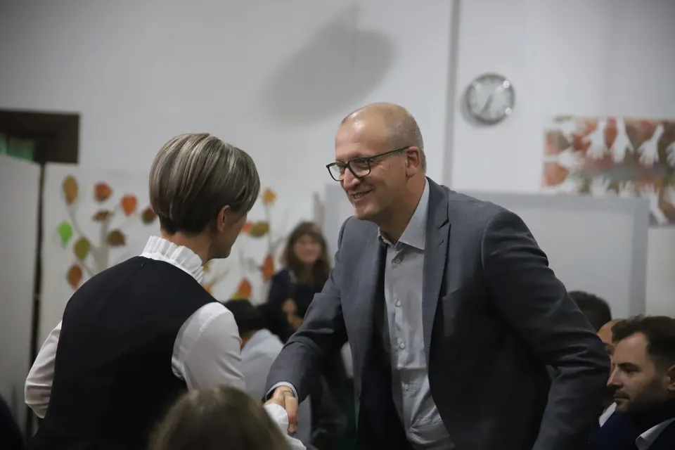 Un bărbat cu ochelari și sacou gri întinde mâna spre o femeie cu vestă neagră și cămașă albă într-o încăpere interioară.