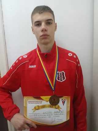 Un tânăr într-o jachetă roșie cu logo-ul JAKO poartă o medalie și ține o diplomă în fața unui fundal alb.