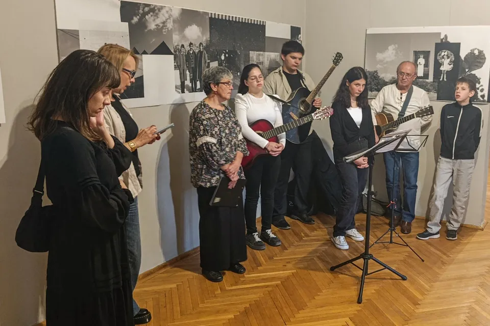 Un grup de persoane stă într-o încăpere cu podea de lemn, în timp ce un bărbat și un tânăr țin chitare acustice.