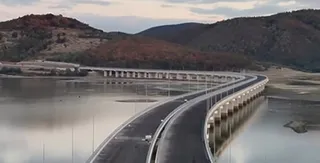 O autostradă cu două caraje curbează pe deasupra unei suprafețe de apă, fiind susținută de piloni albi sub un cer înnorat.