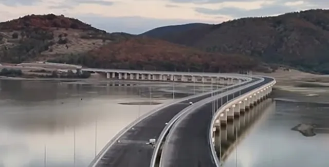 O autostradă cu două caraje curbează pe deasupra unei suprafețe de apă, fiind susținută de piloni albi sub un cer înnorat.