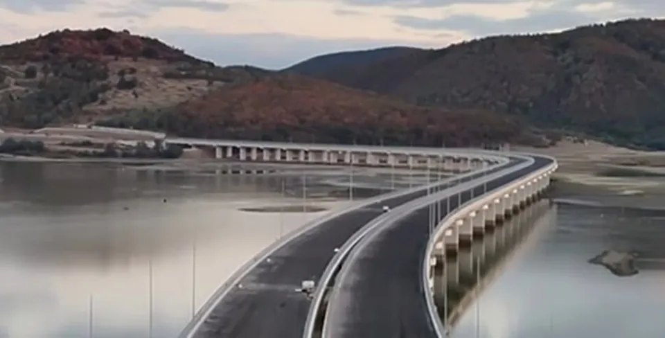 O autostradă cu două caraje curbează pe deasupra unei suprafețe de apă, fiind susținută de piloni albi sub un cer înnorat.