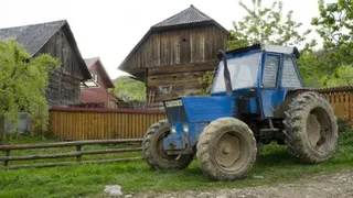 Un tractor albastru este parcat pe un drum de țară lângă niște clădiri din lemn și un gard de lemn.