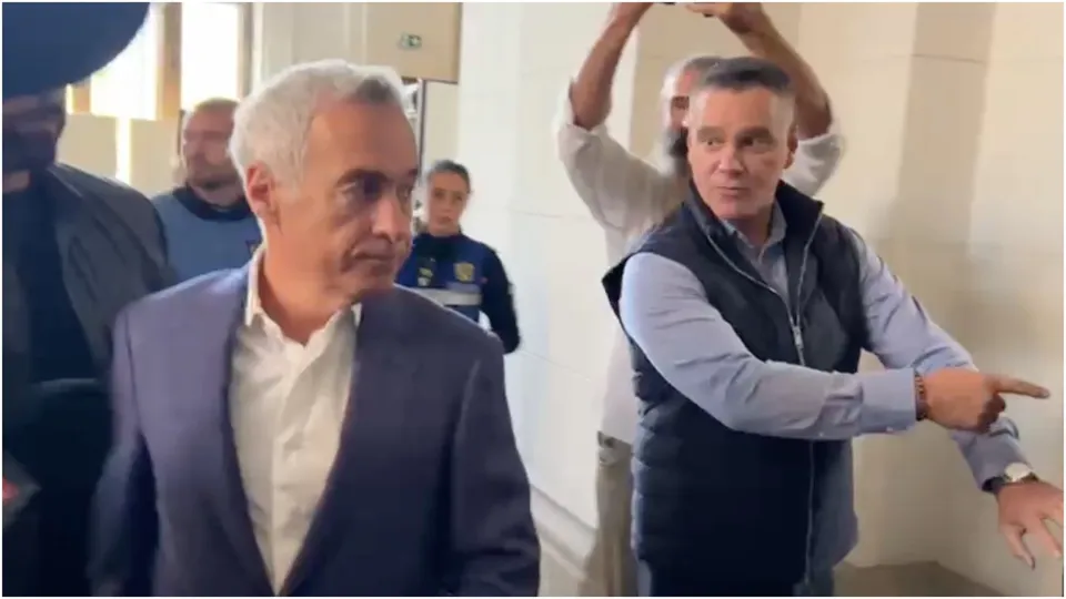 Un bărbat cu părul alb și sacou în prim-plan privește spre lateral, în timp ce un alt bărbat cu vestă și cămașă albastră arată spre dreapta.
