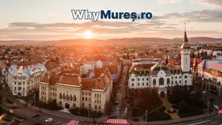 O vedere aeriană a unui oraș la apus, prezentând clădiri cu acoperișuri înclinate, străzi și un turn înalt sub un cer luminos.