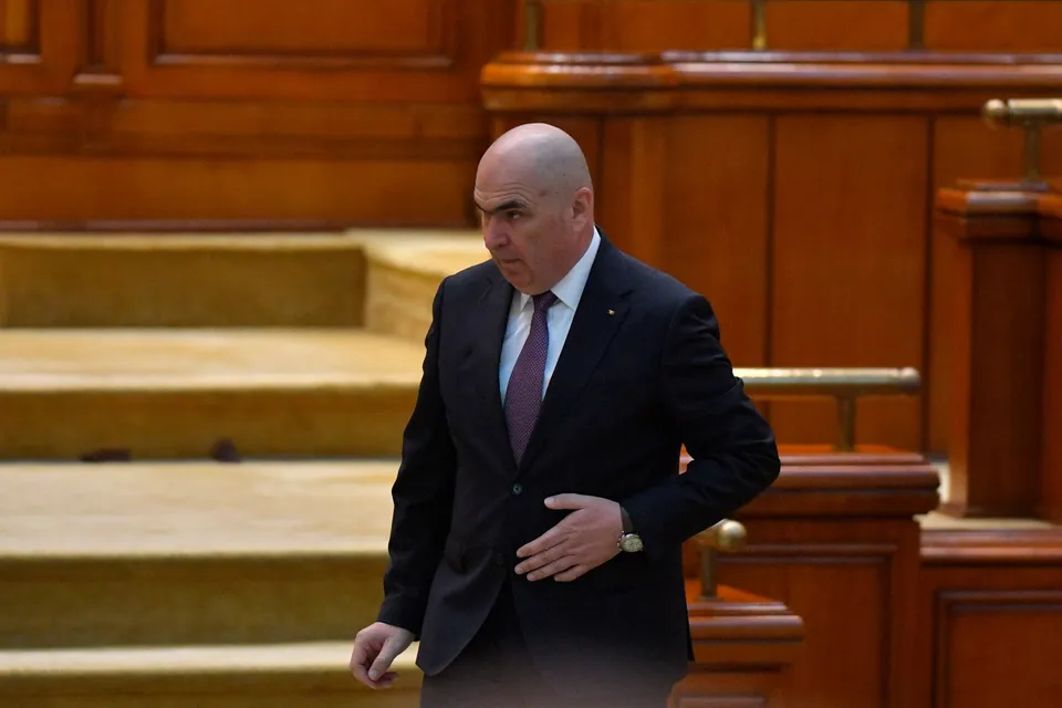 Un bărbat cu capul chel, purtând costum negru, cămașă albă și cravată mov, merge într-un spațiu cu mobilier din lemn și trepte galbene.