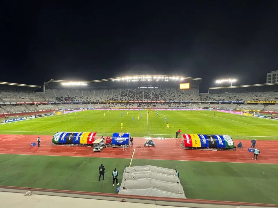 Un teren de fotbal verde este luminat de reflectoare sub un cer de noapte, fiind înconjurat de tribunele unui stadion pline de spectatori.