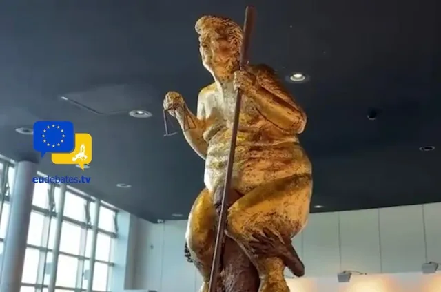O sculptură aurie de o figură umană cu un obiect lung vertical este situată într-o încăpere cu ferestre mari și tavan închis.
