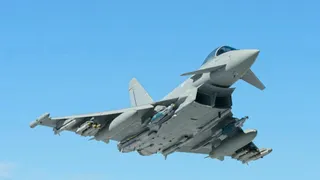 Un avion de luptă gri zboară pe un cer albastru, având diverse rachete și echipamente montate sub aripi.