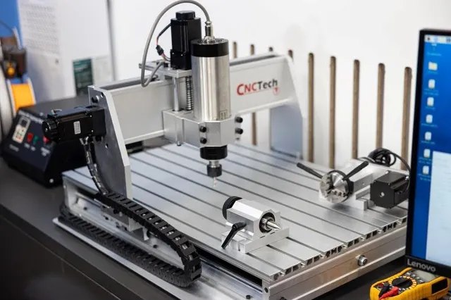 Un aparat CNC cu o suprafață metalică striată, un motor vertical și cabluri negre este poziționat pe o masă lângă un monitor de calculator.