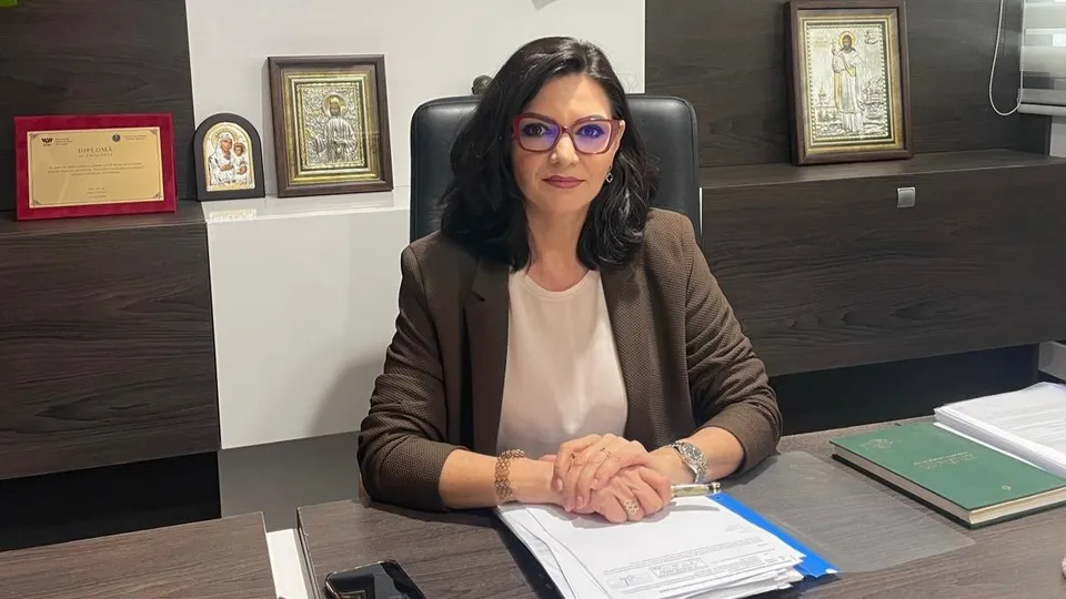 O femeie cu ochelari și sacou maro stă la un birou cu documente, haine albe și cadre în fundal.