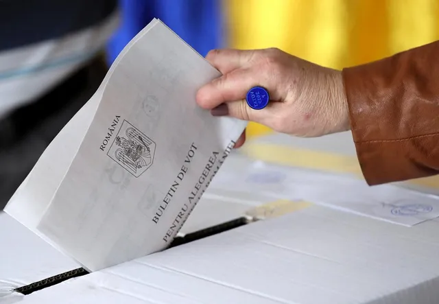 O mână ține un buletin de vot alb cu text și emblemă peste o deschizătură îngustă într-o cutie de vot albă.