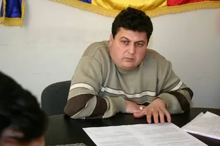 Un bărbat cu părul închis la culoare poartă un pulover maro și bej, stând la o masă cu hârtii în față.