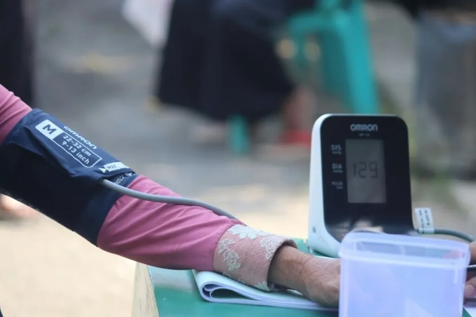 Un braț cu mânecă roz este înfășurat într-un brâțar de tensiometru conectat la un aparat digital care afișează cifra 129.