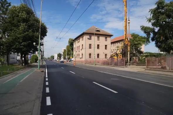 O stradă asfaltată cu linii albe marcaje este desenată lângă un trotuar, un spațiu verde și clădiri cu un utilaj de construcții galben.