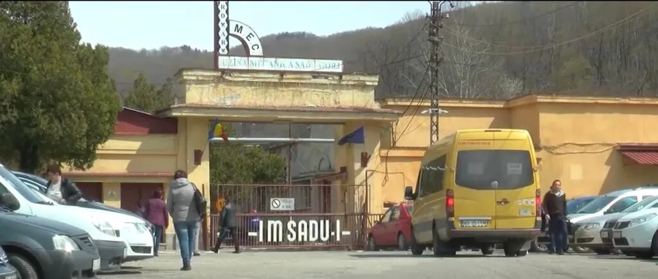 O intrare de fabrică cu un gard metalic, un van galben și mai multe mașini parcate în fața unei clădiri galbene sub un deal împădurit.