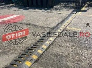 Un dispozitiv de tip bumper cu dungi galbene și negre este montat pe o suprafață asfaltată lângă o marcaj rutier roșu și alb.