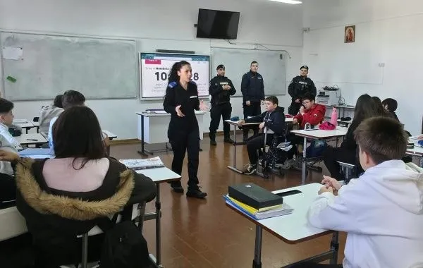 O femeie în uniformă neagră vorbește în fața unor elevi așezați la bănci într-o sală de clasă, în prezența altor persoane în uniformă.