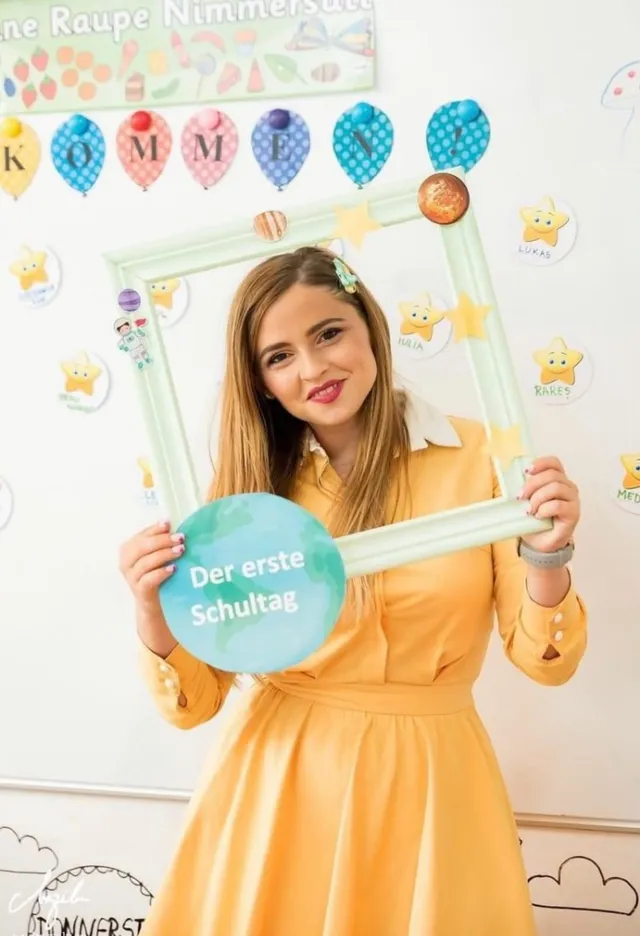 O femeie cu părul lung și rochie galbenă ține un cadru decorativ alb cu un cerc albastru pe care scrie un text în germană.