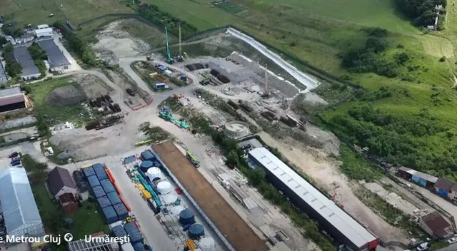 O vedere aeriană prezintă un șantier de construcții cu utilaje, containere colorate, materiale de construcție și drumuri de acces într-o zonă verde.