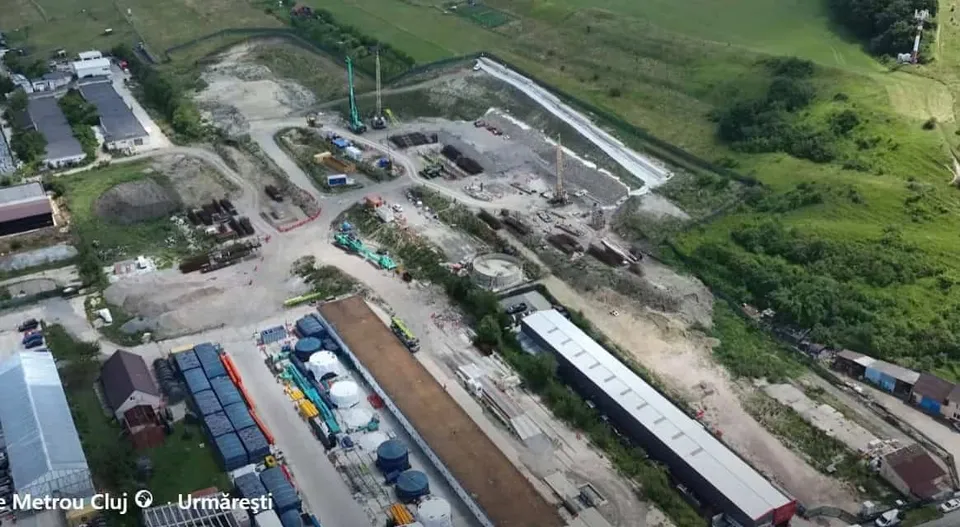 O vedere aeriană prezintă un șantier de construcții cu utilaje, containere colorate, materiale de construcție și drumuri de acces într-o zonă verde.