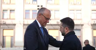 Un bărbat cu ochelari și costum închis își pune mâna pe umărul unui alt bărbat cu barbă, în fața unei clădiri cu ferestre mari.