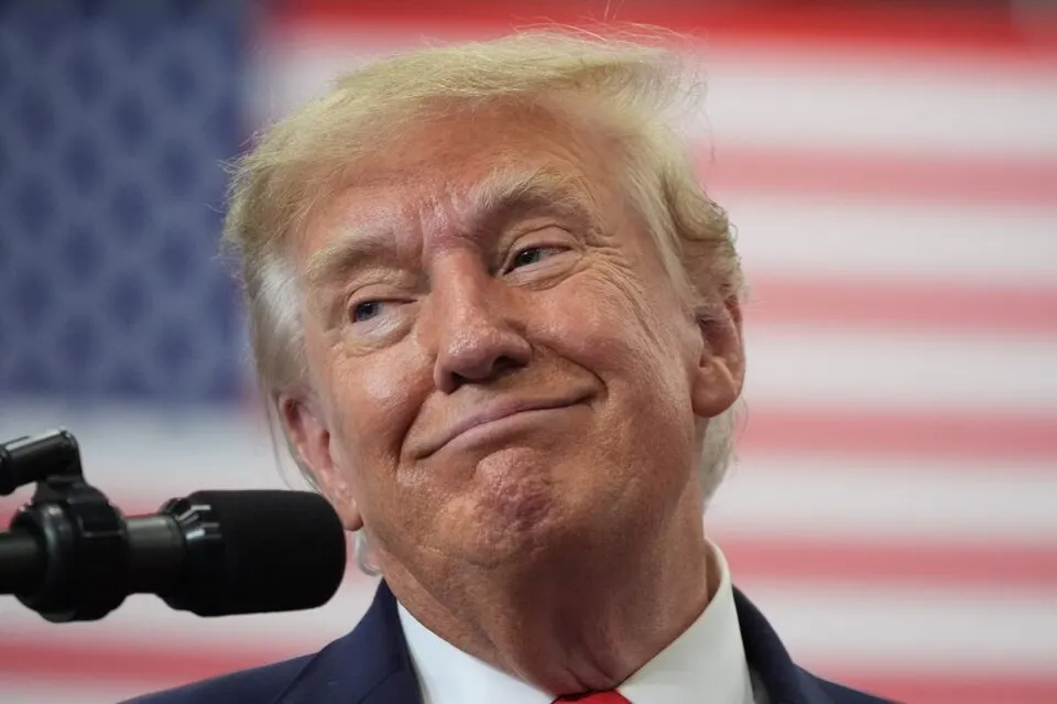 Un bărbat cu păr blond și costum în față unui microfon, având un steag american în fundal.