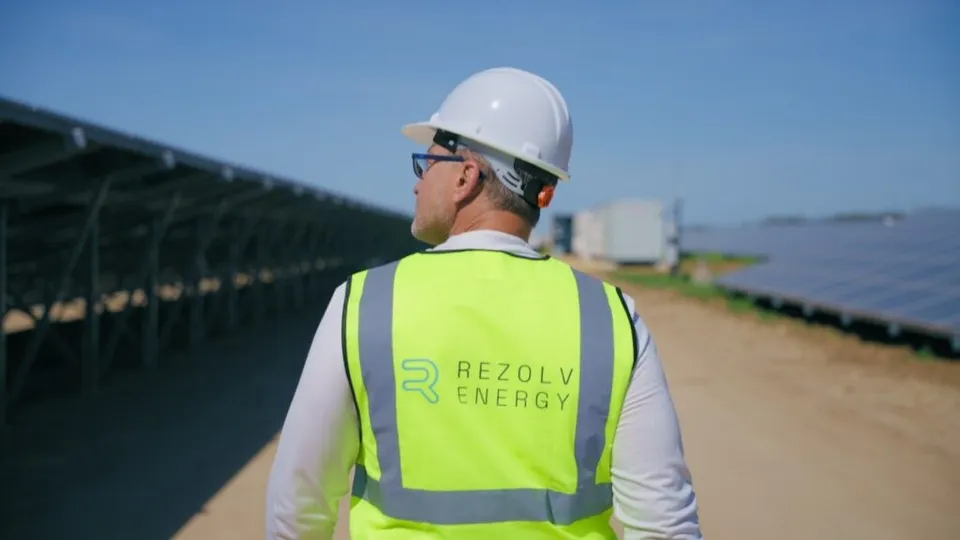 Un bărbat cu cască albă și vestă reflectorizantă cu inscripția Rezolv Energy privește spre rânduri de panouri solare sub un cer senin.