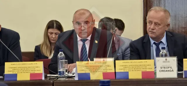 Un bărbat cu ochelari și cravată este așezat la o masă lângă alți bărbați și o femeie, având în față plăcuțe de identificare pe masă.