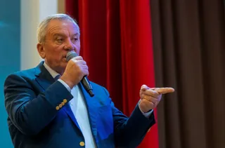 Un bărbat cu părul alb, purtând sacou albastru și cămașă albă, vorbește într-un microfon și arată cu degetul într-o parte.