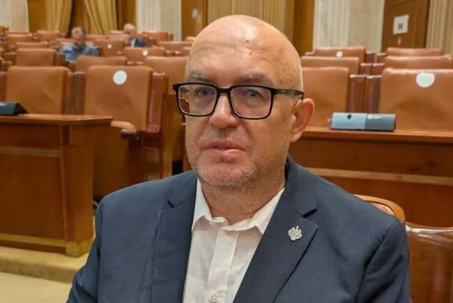 Un bărbat încălțat cu o costum bleumarin, cu ochelari de vedere neîncăadrati, pozitionat într-o sală de ședințe cu bănci de lemn maro.