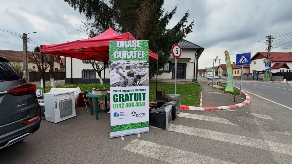 Un afiș vertical verde stă pe trotuar lângă un cort roșu, având în spate un vehicul gri și diverse electrocasnice pe asfalt.