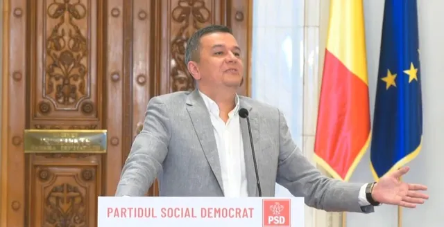 Un bărbat cu sacou gri și cămașă albă stă în fața unor steaguri și unei uși din lemn sculptat.