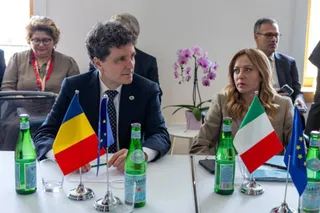 Un bărbat într-un costum închis și o femeie într-o jachetă deschisă stau la o masă cu sticle de apă și steaguri mici.