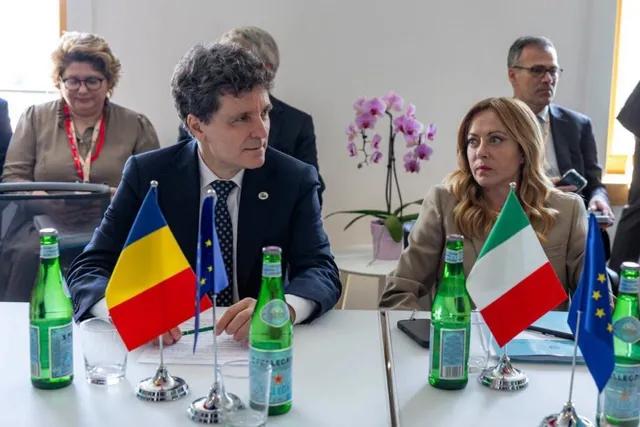 Un bărbat într-un costum închis și o femeie într-o jachetă deschisă stau la o masă cu sticle de apă și steaguri mici.