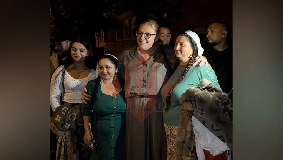 Un grup de femei stă apropiat, unele purtând haine tradiționale sau accesorii precum o batică albă și o coadă împletită.