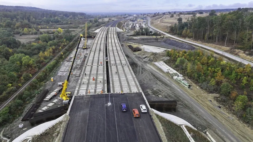 O vedere aeriană prezintă un viaduct în construcție cu mai multe benzi de asfalt, vehicule circulând și utilaje de construcții pe marginea drumului.