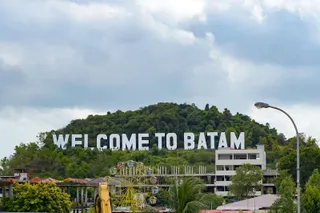 Un semn alb mare cu textul WELCOME TO BATAM este situat pe un deal împădurit sub un cer înnorat.