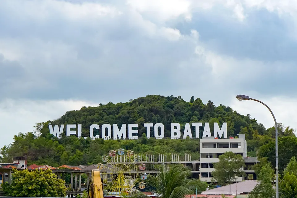 Un semn alb mare cu textul WELCOME TO BATAM este situat pe un deal împădurit sub un cer înnorat.