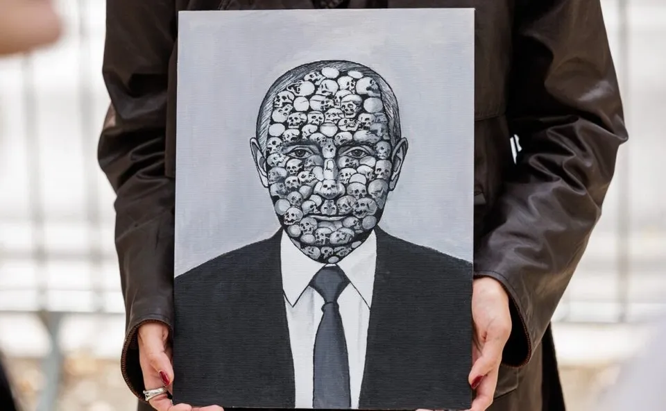 O persoană ține în față o pictură cu chipul lui Putin format din multe fețe, purtând haină închisă la culoare.