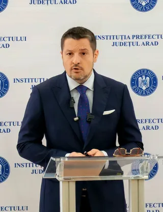 Un bărbat cu sacou închis la culoare și cravată albastră stă în spatele unui pupitru transparent în fața unui fundal alb cu inscripții și embleme.