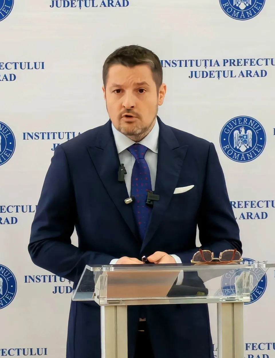 Un bărbat cu sacou închis la culoare și cravată albastră stă în spatele unui pupitru transparent în fața unui fundal alb cu inscripții și embleme.