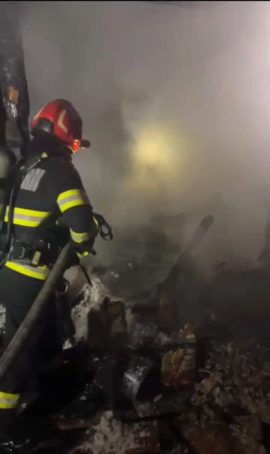 Un pompier cu echipament de protecție și cască este poziționat lângă un furtun, în fața unei zone pline de fum și resturi întunecate.