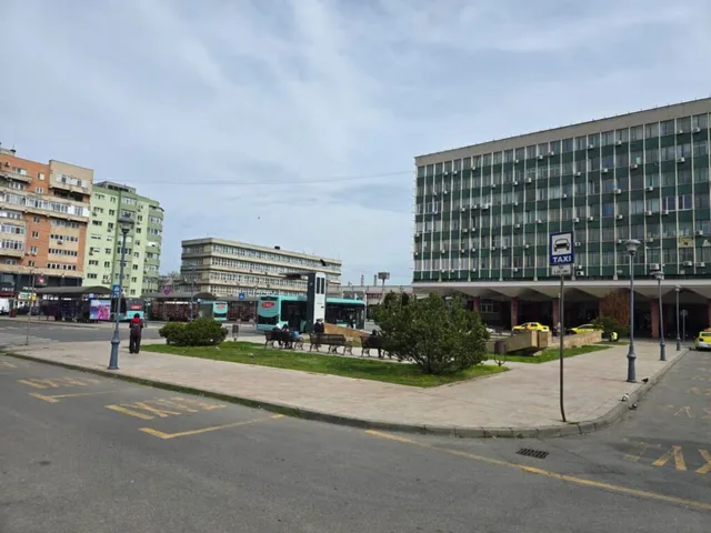 O piață urbană cu clădiri înalte, un spațiu verde cu arbuști și un semn de taxi pe un trotuar gri sub un cer înnorat.