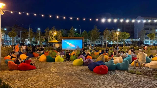 Oamenii stau pe sacoșe colorate pe o piață pavată în fața unui ecran de proiecție mare sub un cer de noapte.